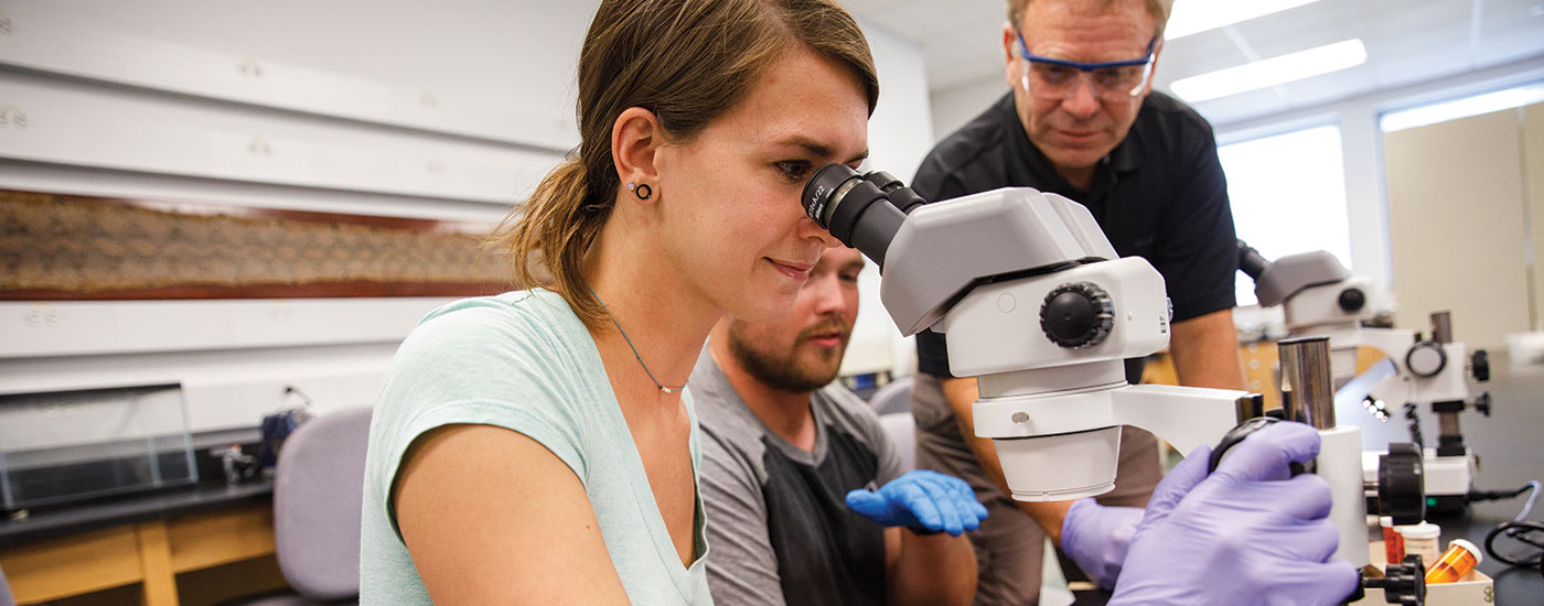 A Frostburg State student looks into a microscope while a professor gives an explanation and another student looks on