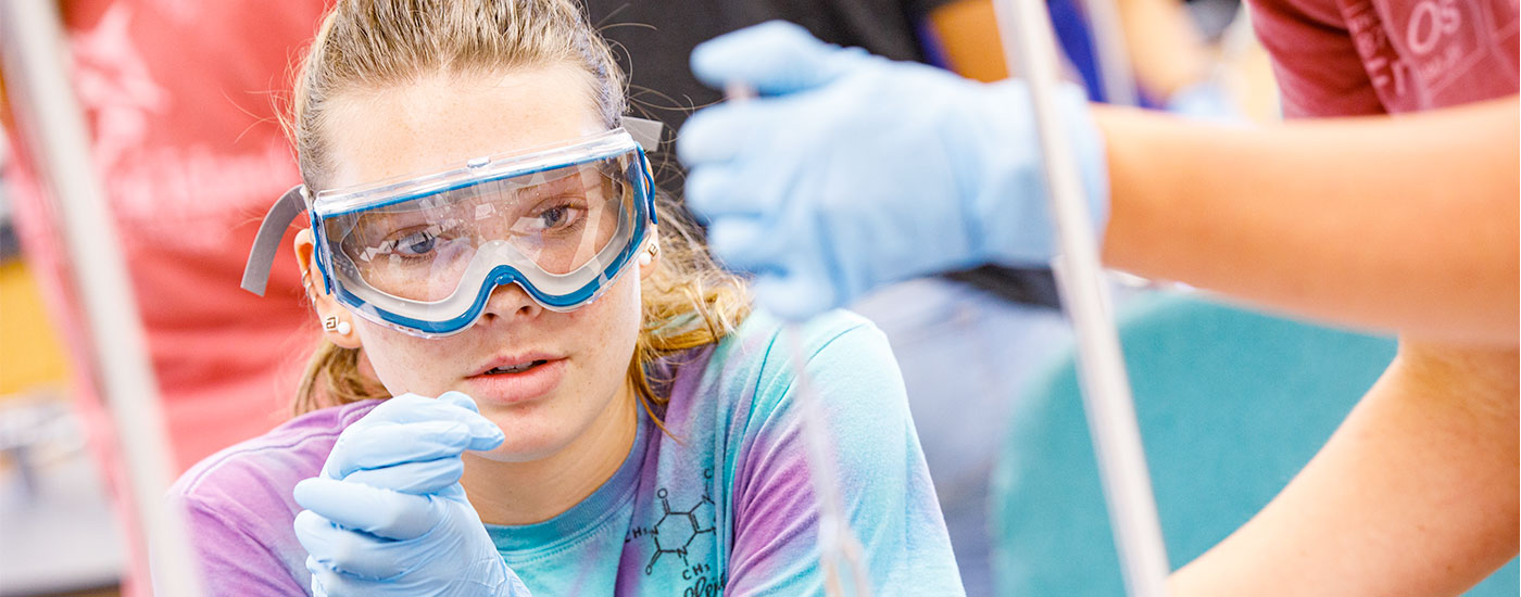 A female Frostburg State student wearing safety goggles observes another student put a liquid in a beaker during a chemistry experiment